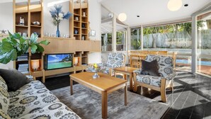 A modern living room with a TV, wooden shelving, patterned furniture, and a view of the outdoors. - 5-bed Haven With Pool Minutes From Beaches (Melbourne)