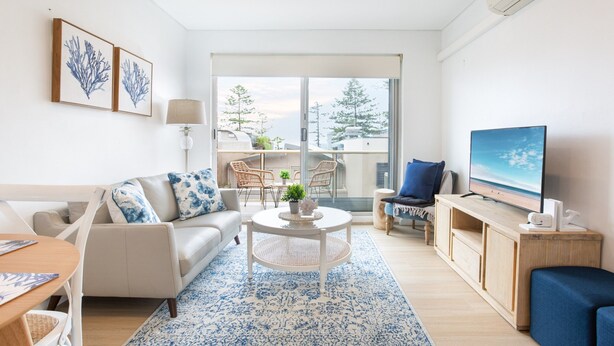 A modern living room with a sofa, coffee table, TV, and a view of a balcony with outdoor furniture.