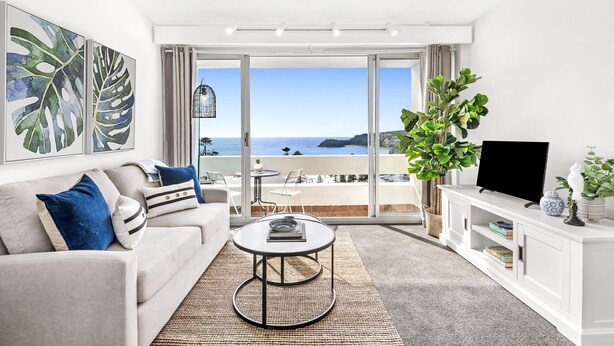 A modern living room with a sofa, coffee table, and a TV on a white console. A large window offers a view of the sea. - Stunning Ocean Views With Manly At Your Doorstep (Sydney)