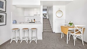 A modern kitchen with white cabinetry, a central island with stools, and a dining area with a wooden table and white chairs. - Stunning Ocean Views With Manly At Your Doorstep (Sydney)