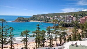 A coastal view with a beach, clear blue sea, and buildings in the background. - Stunning Ocean Views With Manly At Your Doorstep (Sydney)