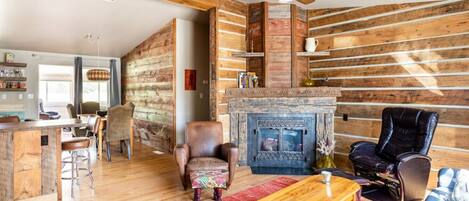 A rustic interior with a wooden log wall, a fireplace, leather armchairs, and a dining area with a bar.
