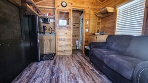 A compact cabin interior with a kitchenette, a wooden floor, a black couch, and a window with blinds. - Christopher Creek Cabins - Cabin 2 (Christopher Creek)