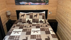 A wooden cabin room with a bed featuring a deer-themed quilt, a painting of a lake, and a nightstand with a lamp. - Christopher Creek Cabins - Cabin 2 (Christopher Creek)