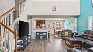 Un salón amplio con una escalera de madera, un televisor de pantalla plana, una mesa de comedor con sillas, un sofá de cuero y una estantería.