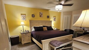 Un dormitorio con cama, mesitas de noche, un banco, ventilador de techo y una puerta.