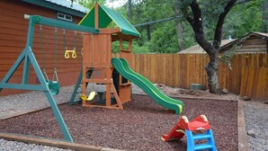 Une structure de jeux en bois, dotée d’un toboggan et de balançoires, entourée de gravier et d’arbres.