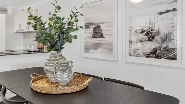 Un comedor con una mesa de madera oscura, un florero a rayas con follaje verde y dos cuadros en blanco y negro de paisajes costeros en la pared.