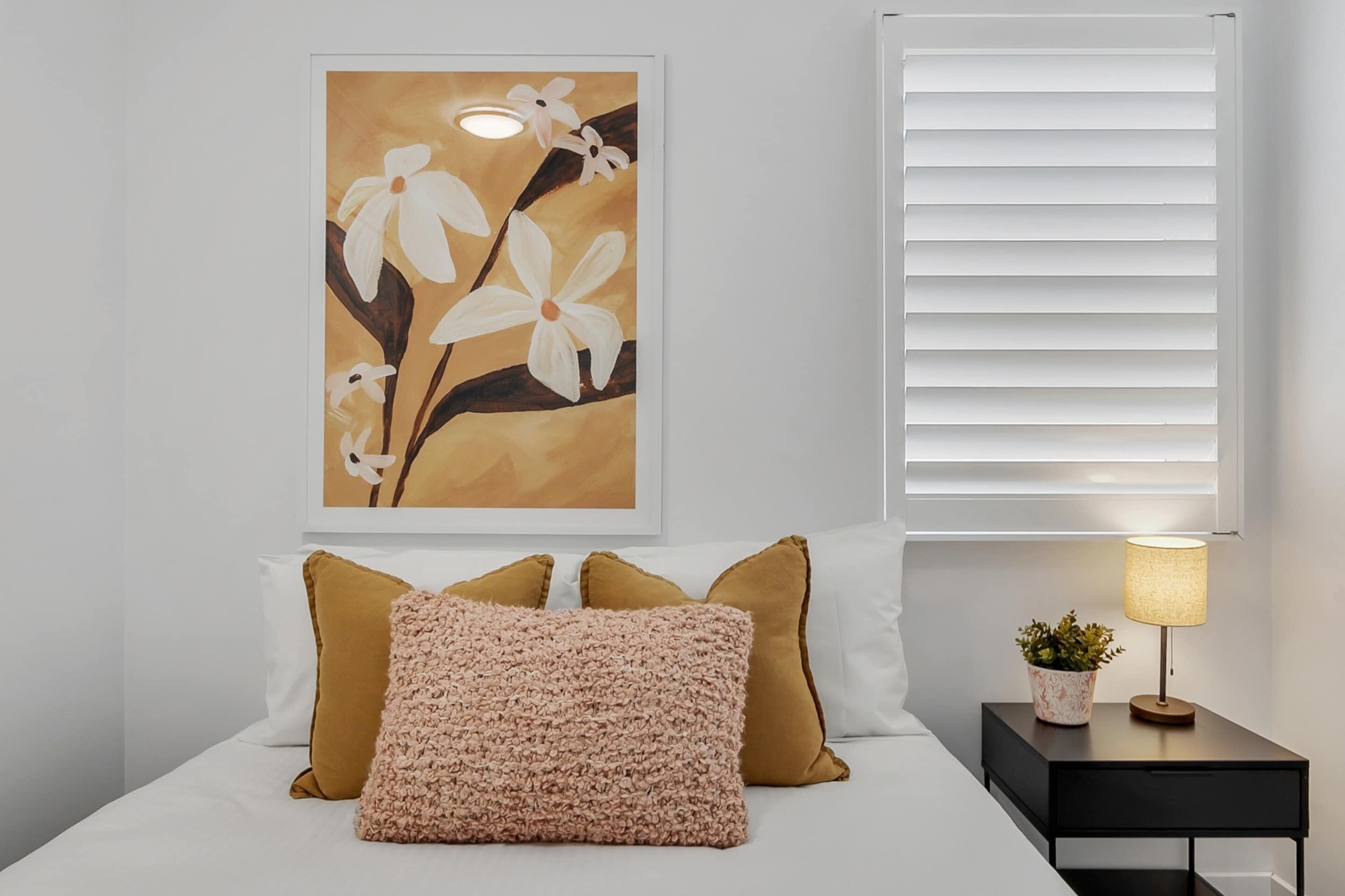 A bedroom with a bed, pillows, a nightstand with a lamp, a wall art of flowers, and a window with blinds.