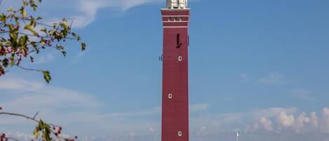 Ein hoher, roter Leuchtturm mit Wetterfahne, umgeben von Sträuchern und einem wolkenlosen Himmel.