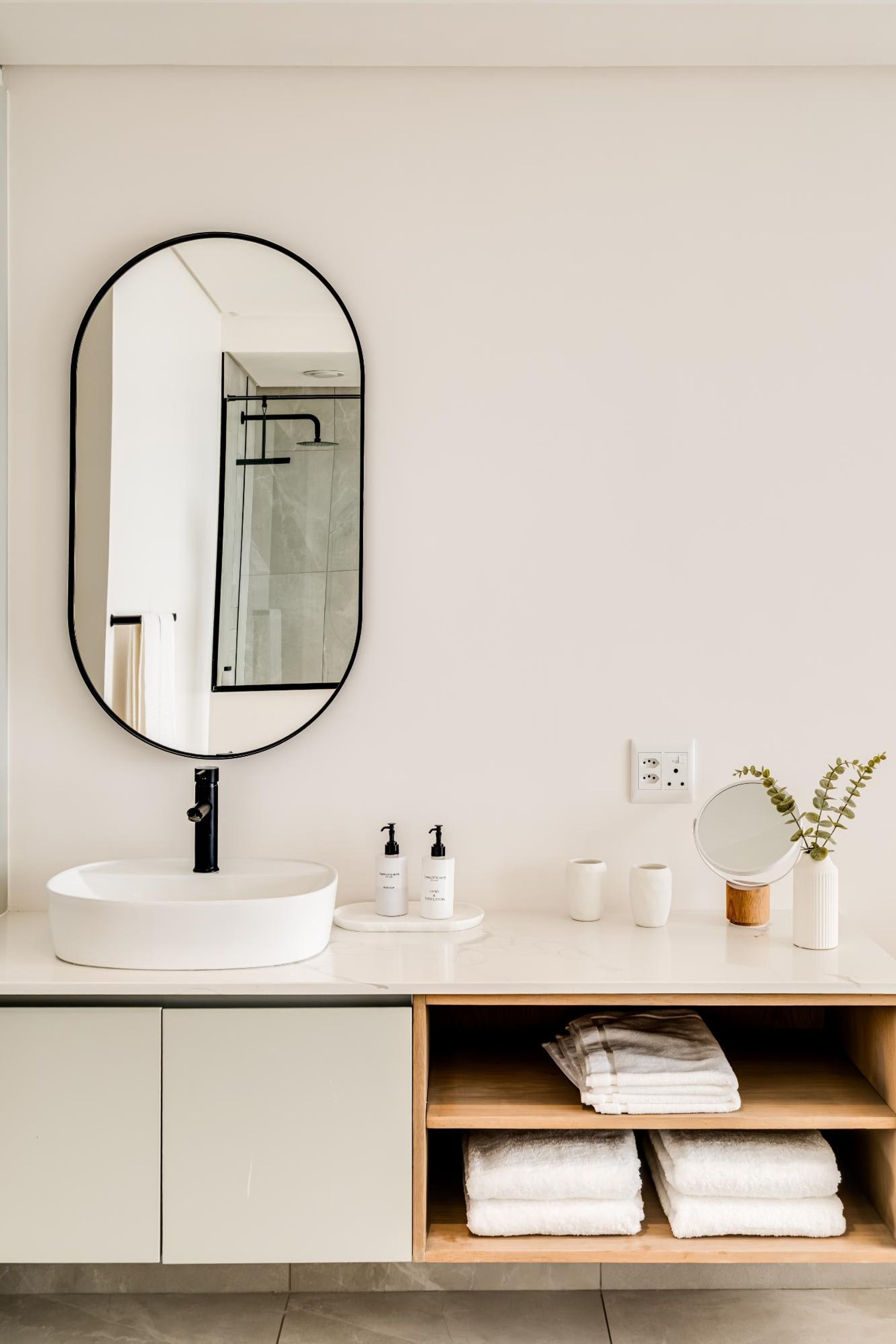 A modern bathroom with a round mirror, white sink, and a wooden shelf with towels.