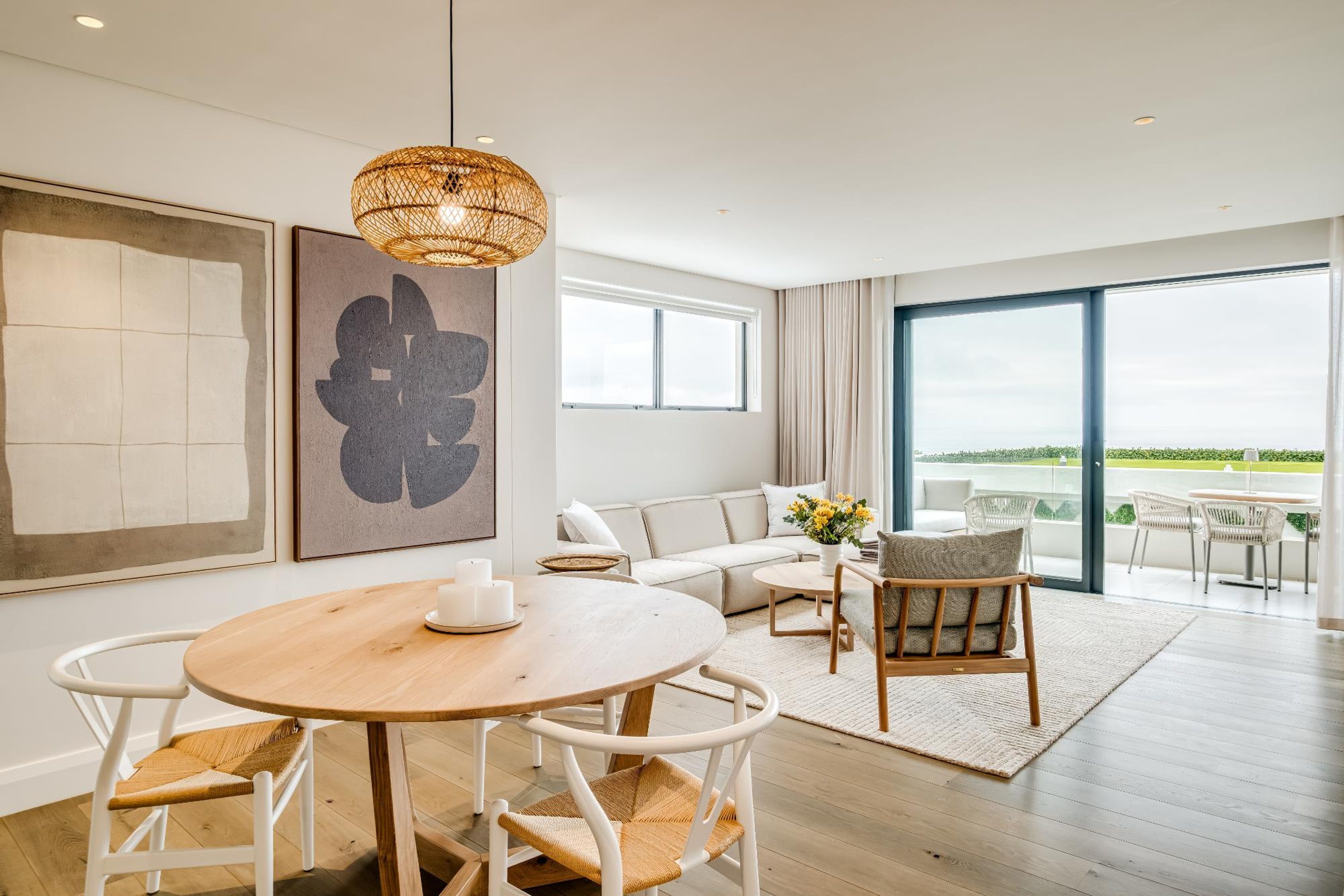 A modern living room with a round wooden table, white chairs, a sofa, and a large window offering a view of the outdoors.