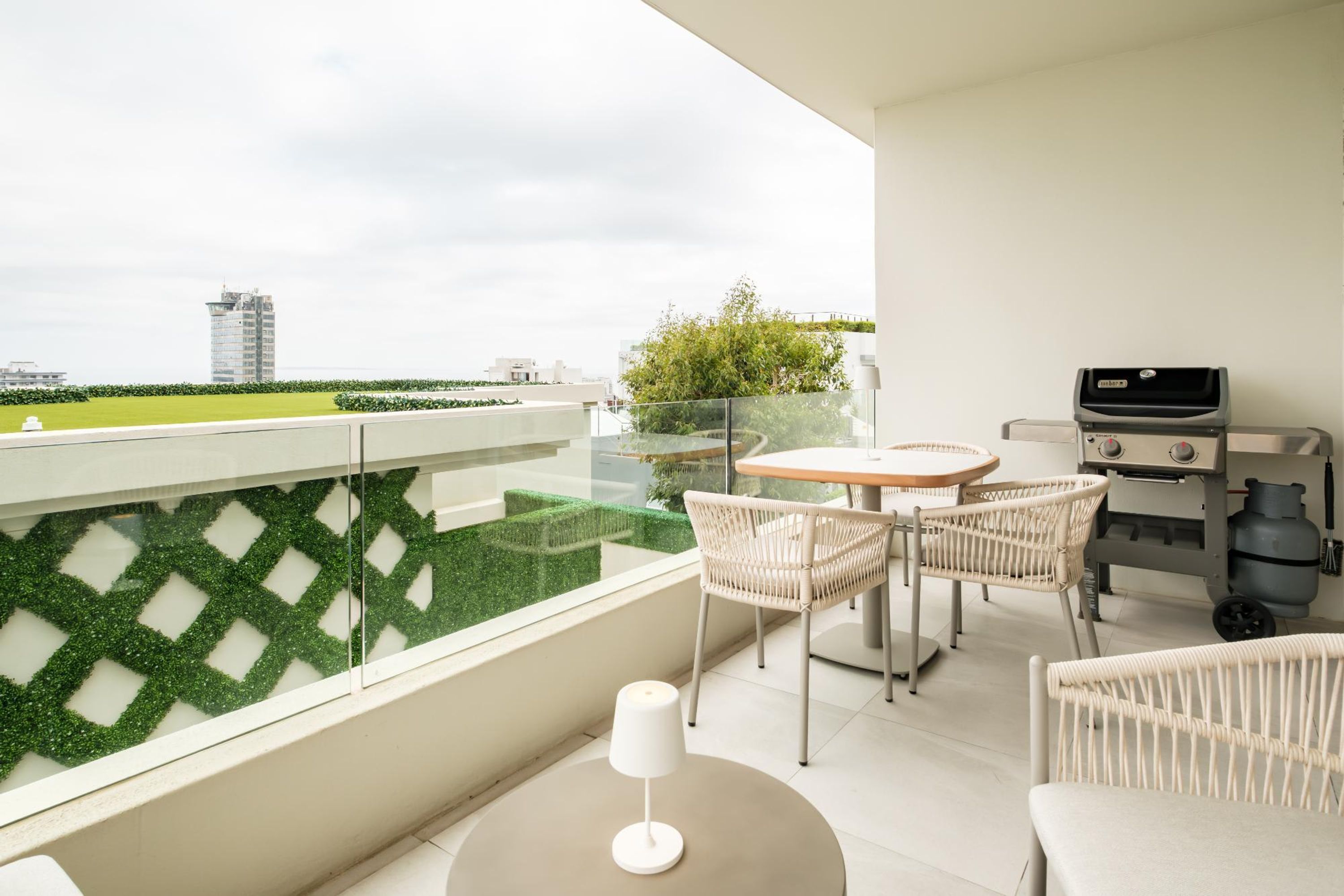 A balcony with a glass railing, a table with chairs, a grill, and a view of a green field and buildings.