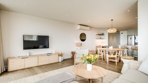 Un salon moderne doté d’un téléviseur à écran plat, de meubles en bois, d’un coin repas avec une table et des chaises en bois, et d’une cuisine en arrière-plan.