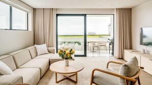 Un salon moderne avec un canapé, un fauteuil et une table basse. Un téléviseur est fixé au mur, et un balcon avec des meubles d’extérieur est visible à travers les portes vitrées.