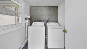 Une buanderie équipée de deux machines à laver blanches, d’un sèche-linge et d’une fenêtre avec des stores.