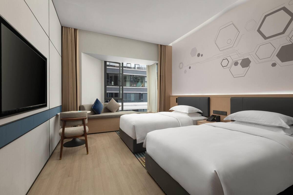 Une chambre d’hôtel avec deux lits, une télévision, un bureau et une fenêtre donnant sur un immeuble.