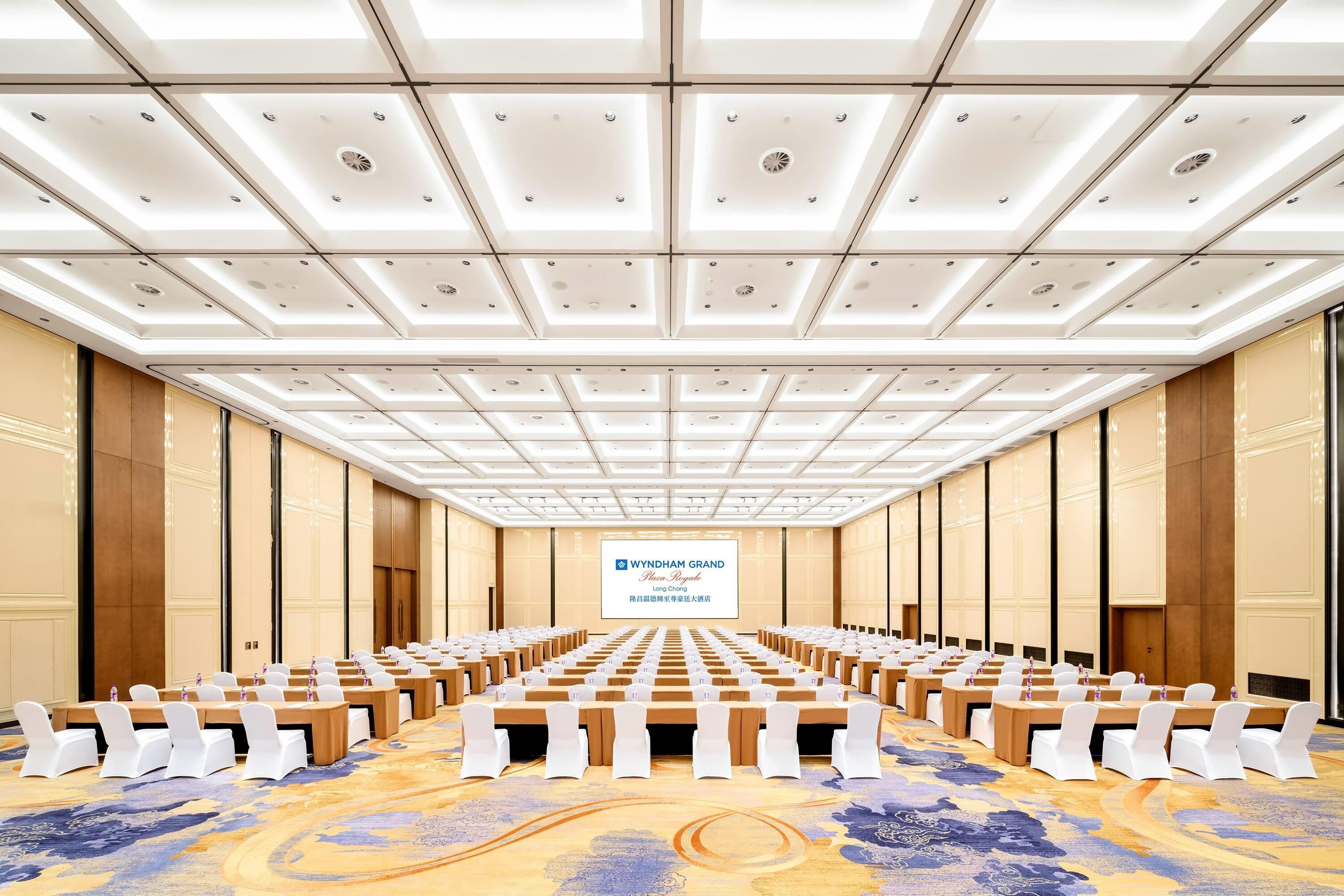 Un salón de conferencias con filas de mesas y sillas, una pantalla grande al frente y una alfombra estampada.