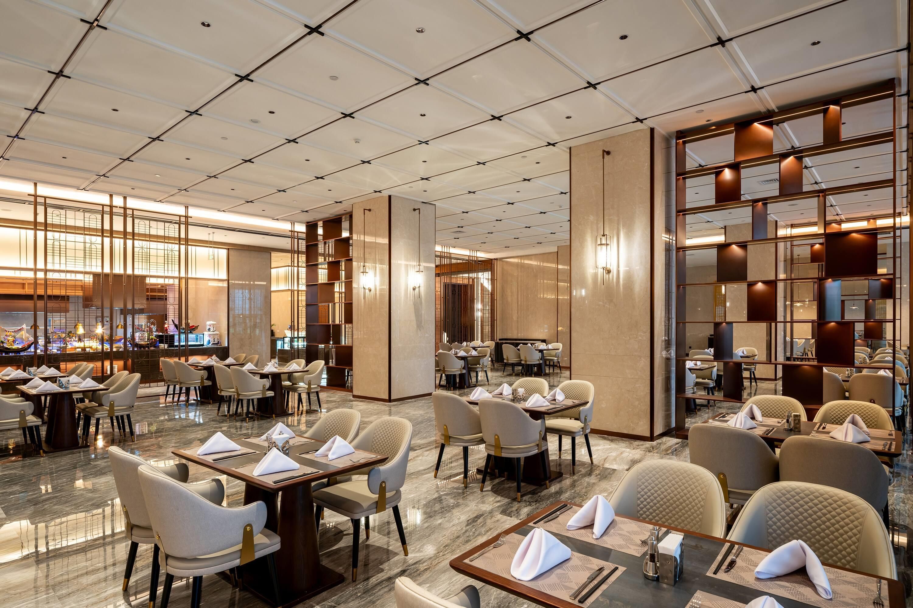 A modern hotel dining area with multiple tables and chairs, a kitchen in the background, and a high ceiling with a grid pattern.