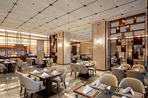 Un espace de restauration moderne dans un hôtel, avec de nombreuses tables et chaises, une cuisine en arrière-plan et un plafond haut orné d’un motif en réseau.
