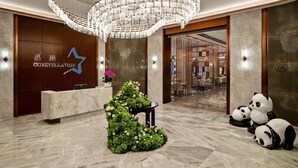 Un área de recepción de hotel con diseño moderno, que incluye una gran lámpara de araña, un mostrador de recepción, una decoración con plantas y un cartel con el nombre del hotel.