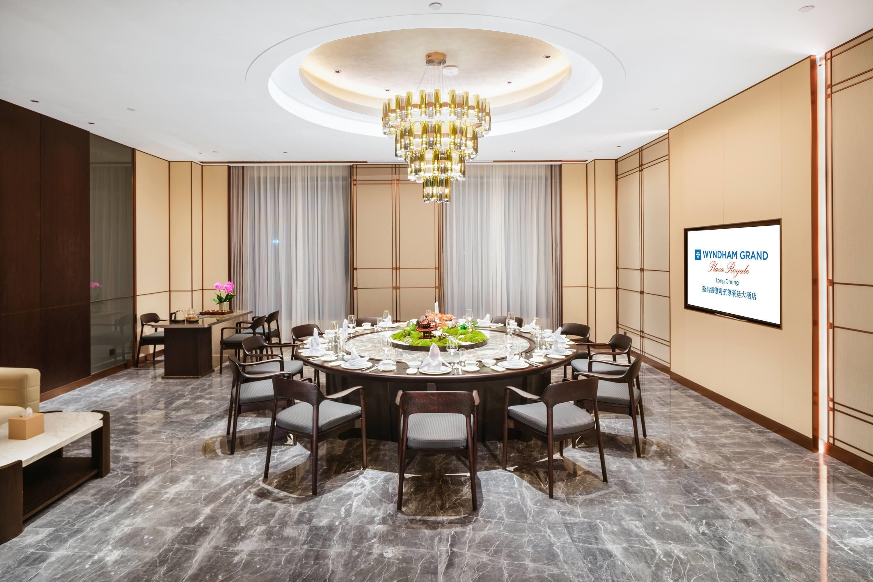Un salón de conferencias con una mesa redonda dispuesta para una reunión, una araña y una pantalla de proyección mostrando información del hotel.