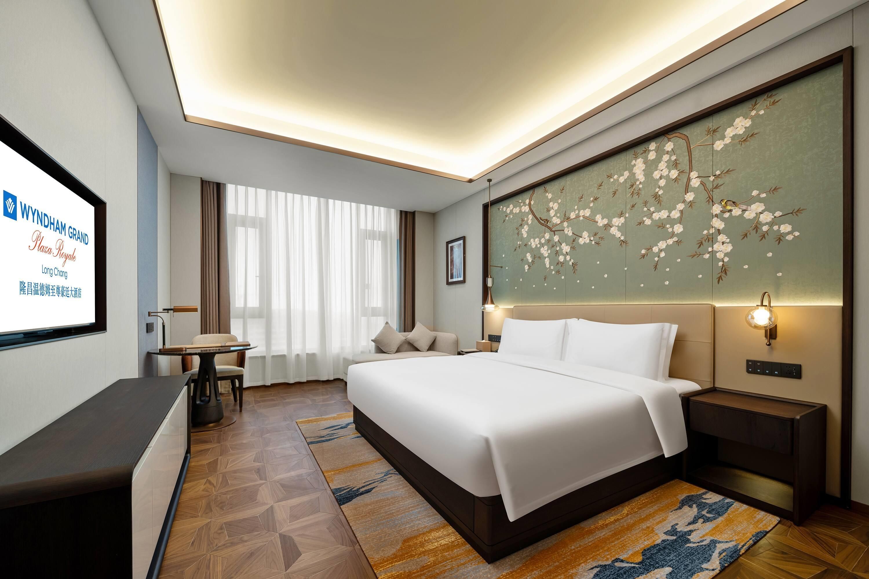 Una habitación de hotel con cama, mesitas de noche, escritorio y una pantalla grande que muestra información del hotel.