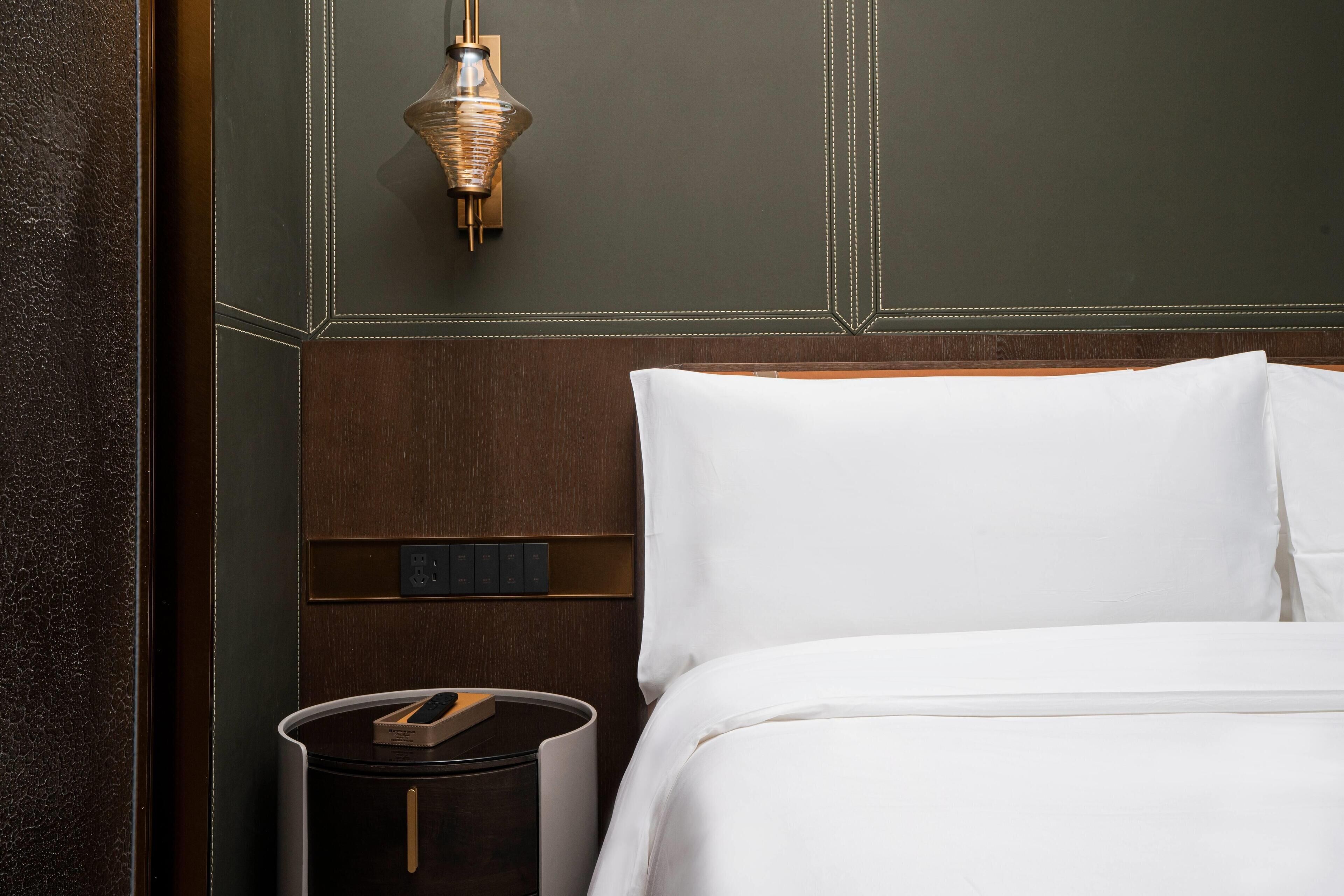 Een hotelkamer met een houten hoofdbord, een nachtkastje met een lamp en een bedieningspaneel aan de muur.