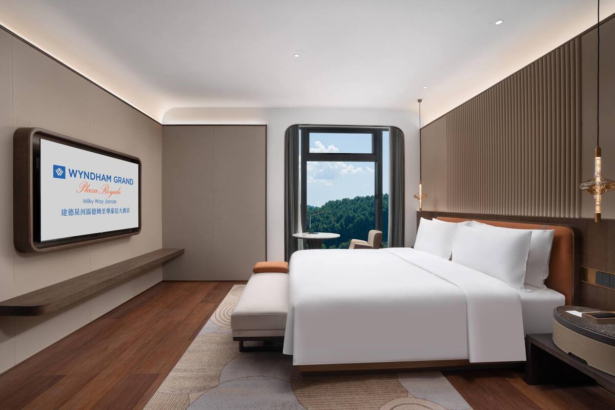 A modern hotel room with a large bed, a mounted TV displaying hotel information, a small round table, and a view of the outdoors.