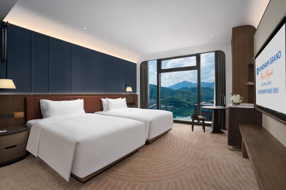 A hotel room with two beds, a desk, a chair, and a large window offering a mountain view.
