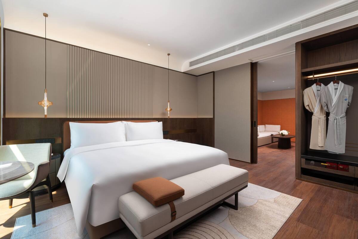 A modern hotel room with a large bed, a seating area, a vanity with a mirror, and a walk-in closet with robes.