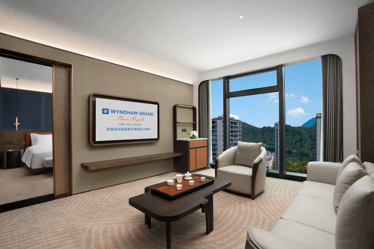 A modern hotel room with a large bed, a sitting area with a sofa and armchairs, a coffee table, and a view of a cityscape through large windows.