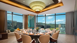 A dining area with a large round table set for eight, surrounded by chairs, with a view of a cityscape through large windows.