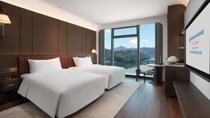 A modern hotel room with two beds, a large window offering a mountain view, a small table with a vase of flowers, and a wall-mounted screen displaying the hotel name.
