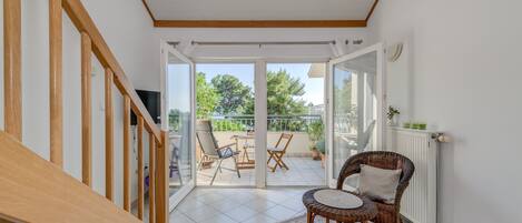 A room with a wooden staircase, a wicker chair, a small table, and a view of an outdoor balcony with chairs and plants.