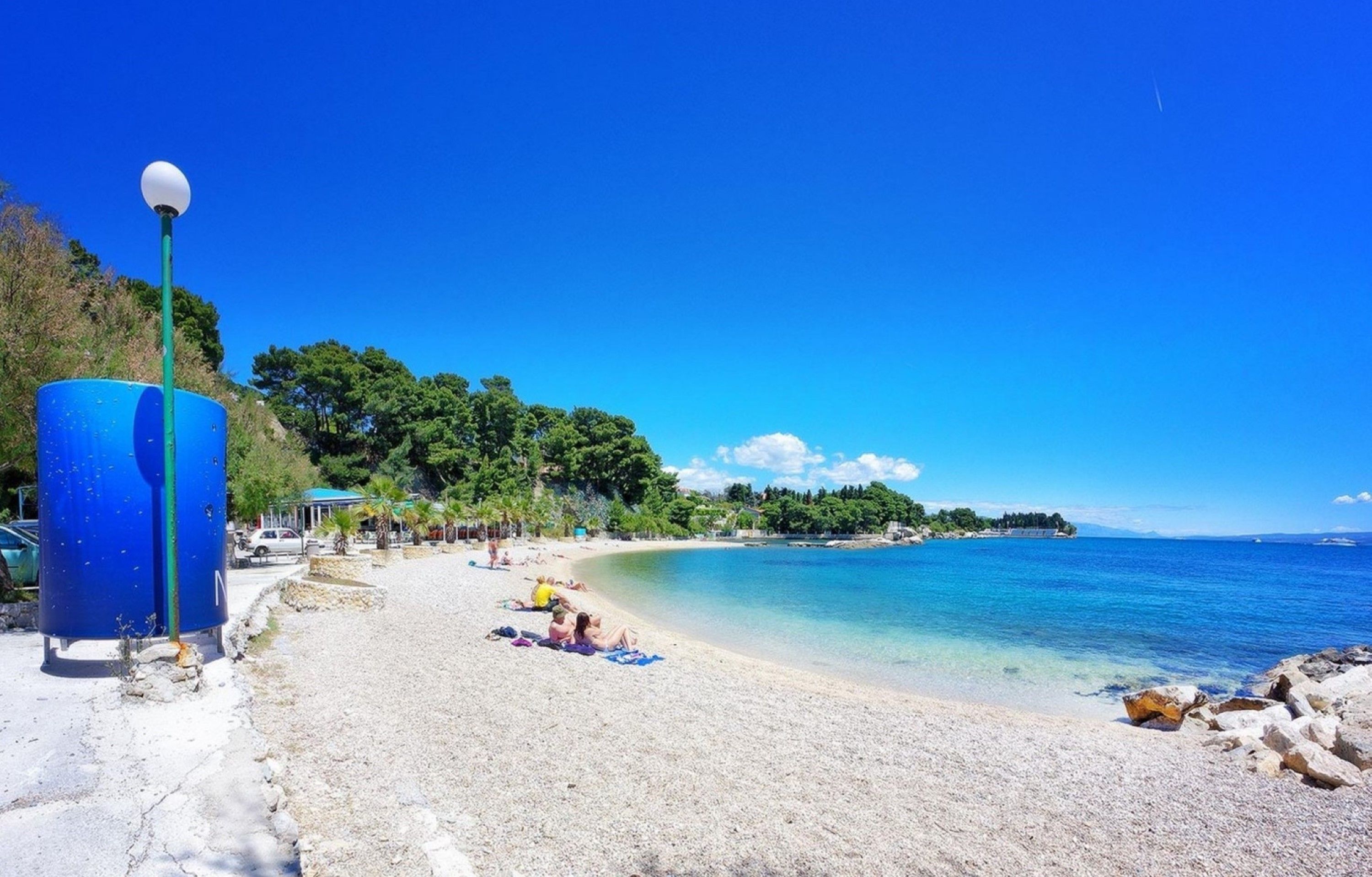 A beach with clear blue water, a few people sunbathing, and a blue lamp post.