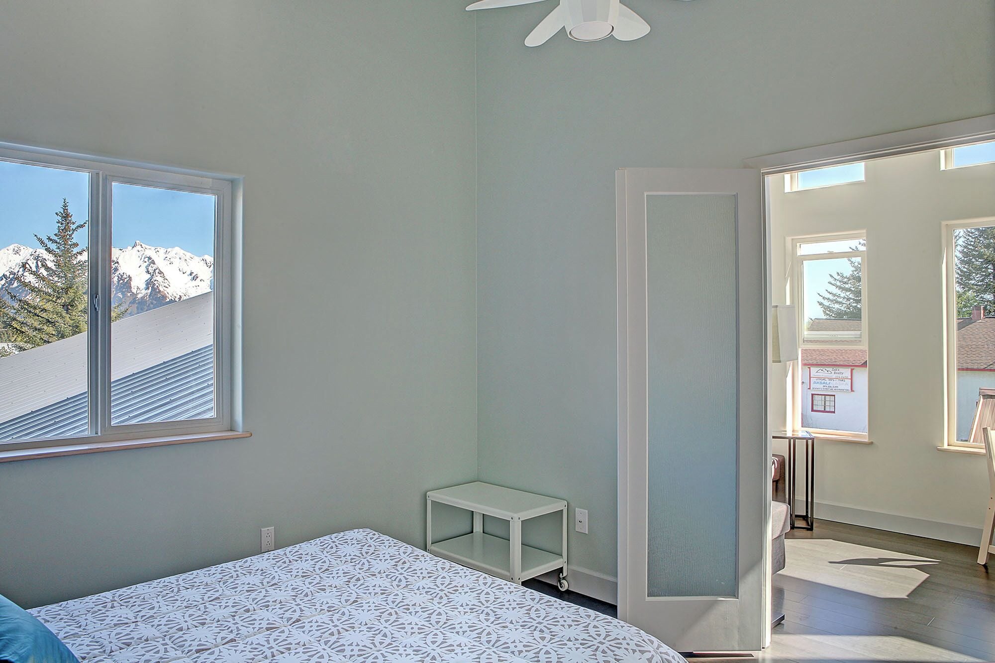 Un dormitorio con una cama, una mesita de noche pequeña y blanca, una ventana con vista a la montaña y una puerta que da a otra habitación.