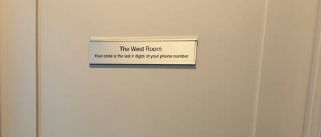 Uma porta com fechadura eletrônica e uma placa acima com a inscrição 'The West Room' que fornece um código baseado nas últimas quatro dezenas de um número de telefone.