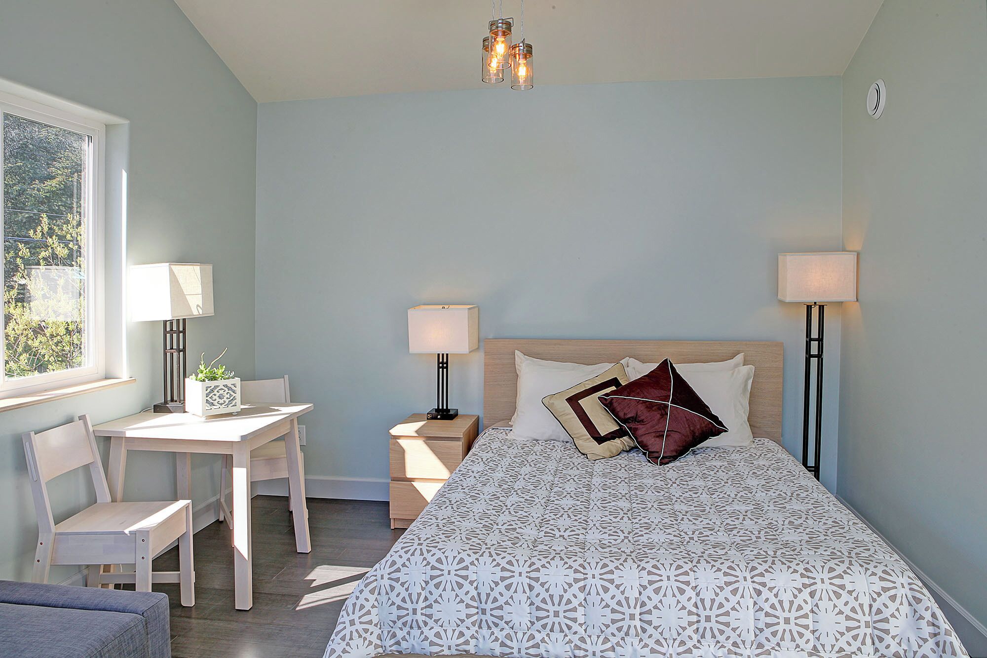 A bedroom with a bed, two bedside tables with lamps, a desk with a chair, and a window with a view of trees.