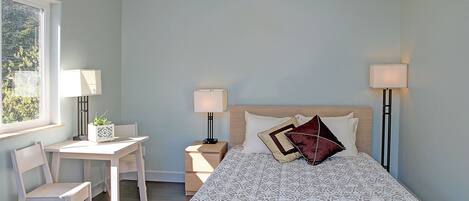 A bedroom with a bed, two bedside tables with lamps, a desk with a chair, and a window with a view of trees.