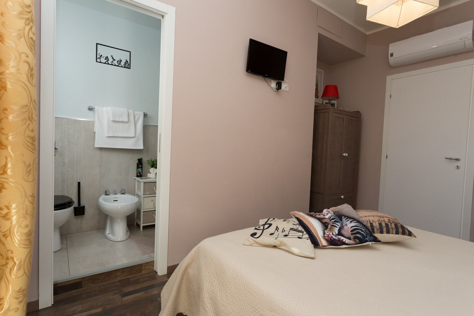 Quarto com cama, almofadas, televisão, guarda-roupa e banheiro visível por uma porta aberta.
