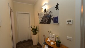 A hallway with a door, a potted plant, a wall-mounted light, a framed picture, a small table with a plant, and a thermostat.