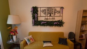 A yellow sofa with two pillows, a wall-mounted decorative piece with a tree design, a small table with a lamp and flowers, and a bookshelf.