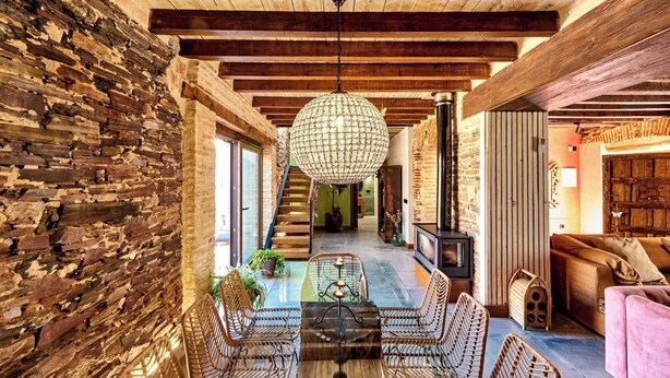 A rustic interior with exposed brick walls, a wooden staircase, a glass dining table, wicker chairs, and a cozy seating area with a pink sofa. - El Nacimiento Santa Elena by Ruralidays (Jaén)