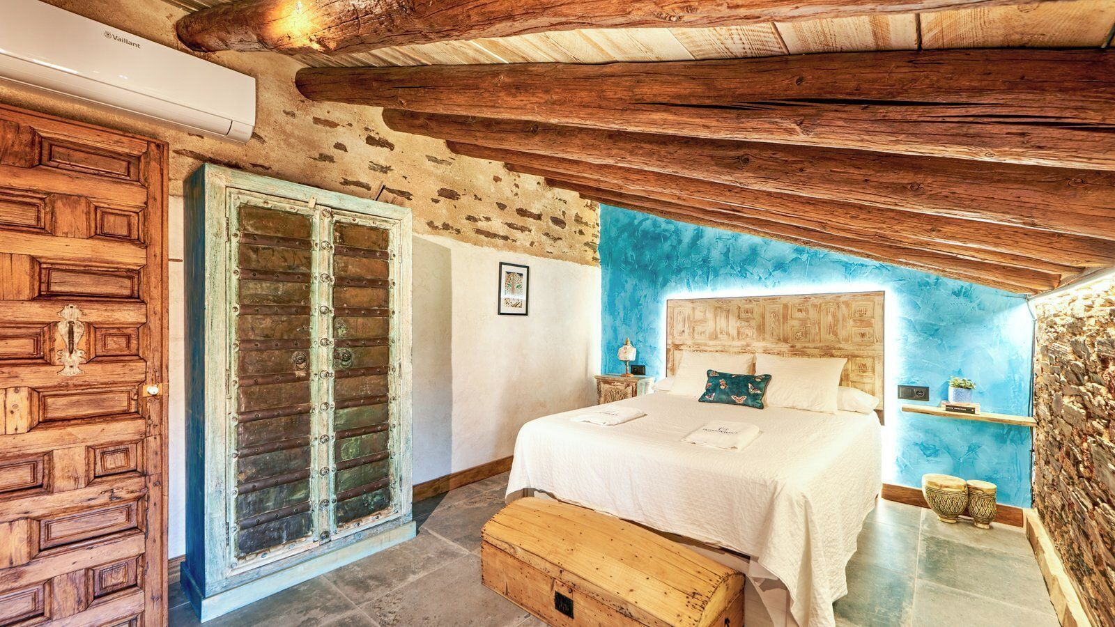 Un dormitorio con techo de madera, una cama con ropa de cama blanca, un cofre de madera y una pared azul de acento.