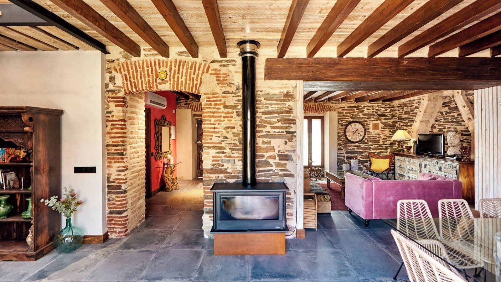 A rustic interior with exposed brick walls, a stone fireplace, wooden beams, and a cozy seating area with a pink sofa and wicker chairs.