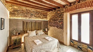 A bedroom with a stone wall, wooden beams, a bed with a floral bedspread, a nightstand, and a vase with flowers. - El Nacimiento Santa Elena by Ruralidays (Jaén)