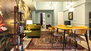 Een moderne woonkamer met een groene bank, een eettafel met stoelen, een kamerkleed met patroon en een boekenplank met boeken en decoratieve objecten.