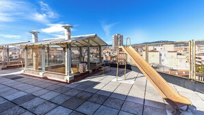 Een terras op het dak met glazen omheining, een houten glijbaan en uitzicht over de stad.