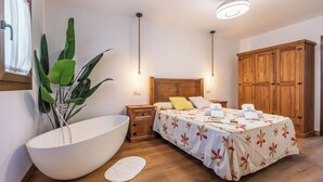 A bedroom with a large white bathtub, a wooden wardrobe, a bed with a floral bedspread, and a potted plant.